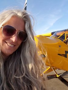 A woman with long grey hair and sunglasses next to an airplane.